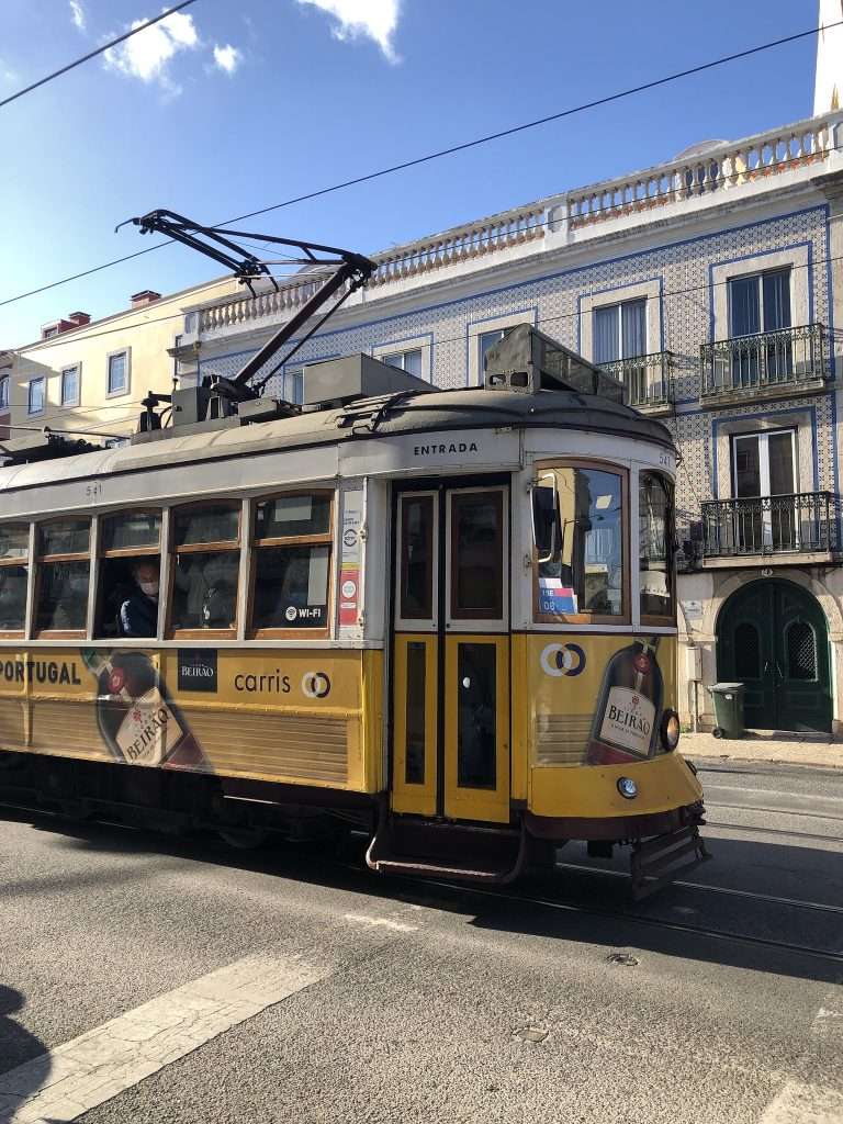 In this photo, we see one of Lisbon's famous trams, a beloved and iconic symbol of the city. The yellow tram stands out against the tiled buildings of the city, its vintage design a testament to the city's rich history and cultural heritage. The tram looks like it's just starting its journey, with passengers visible through its windows, ready to take in the sights and sounds of Lisbon as they travel through the city's narrow, winding streets. The tram tracks stretch out in front of the tram, a reminder of the city's well-developed public transportation system that makes it easy to explore all that Lisbon has to offer. Overall, this photo perfectly captures the charm and character of Lisbon, inviting us to come and experience all that this amazing city has to offer