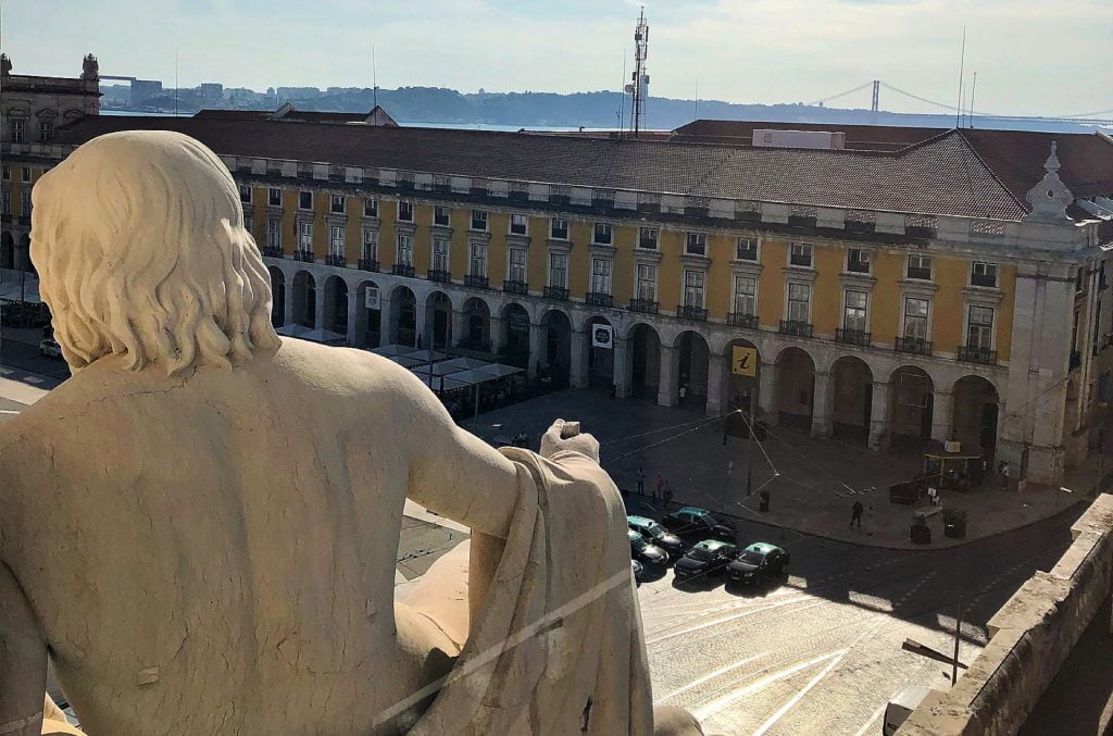 Statue's back view overlooking a historic square in Lisbon with the Rua Augusta Arch in the distance.
