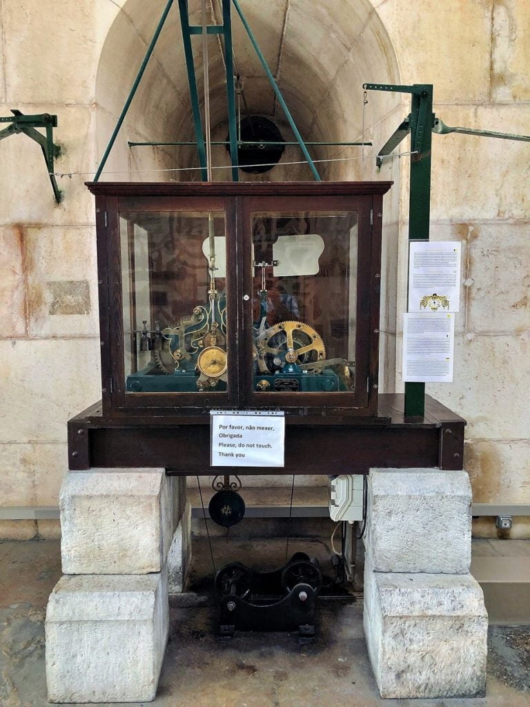 A vintage clock mechanism displayed in a glass cabinet with instructions in Portuguese and English in Lisbon.