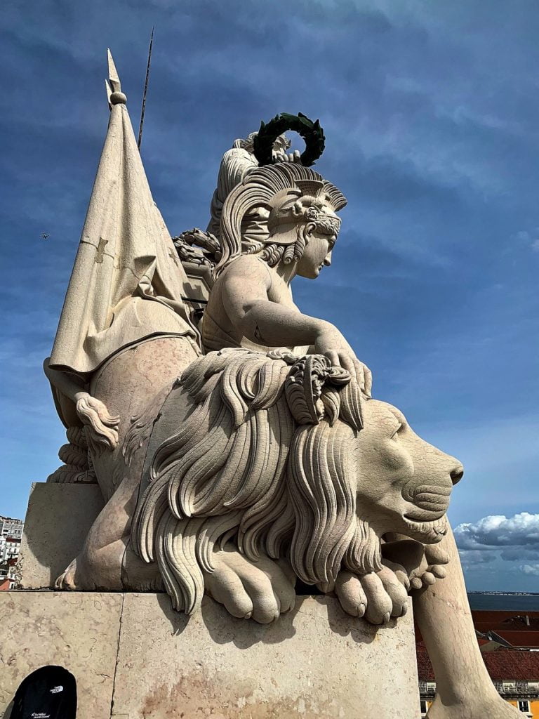 Majestic statue featuring a woman and a lion, with intricate details, set against a blue sky backdrop, at the Rua Augusta Arch in Lisbon.