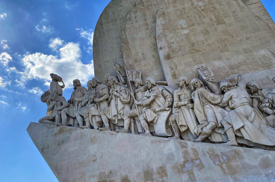 Statues of The Monument to the Discoveries set against a blue sky with clouds.