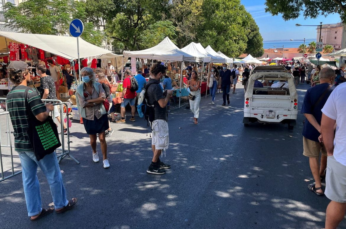 A bustling outdoor flea market on a sunny day with shoppers browsing stalls under white tents, and a small Piaggio truck parked on the street