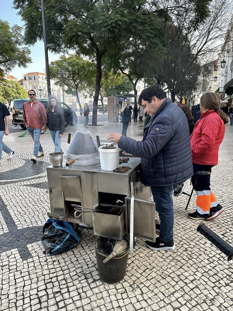 A vendor in a blue jacket serves chestnuts from a street cart in a busy public square in Lisbon Portugal lined with trees and historic buildings in the background.