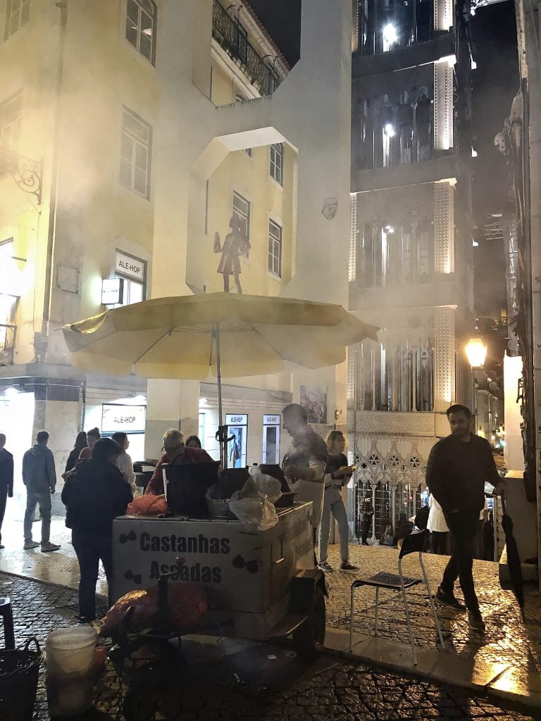 A bustling evening street scene in Lisbon Portugal featuring a chestnut vendor under a yellow umbrella, with the steam from the roasting adding a mystical quality to the surroundings of historical buildings and a mural.